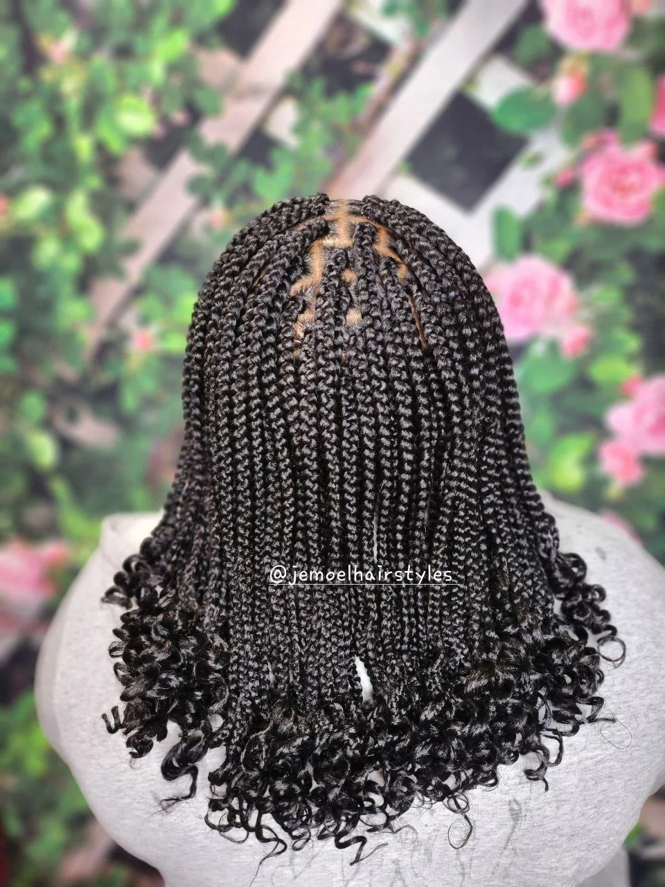 Back view of small boho braids with a middle part, ending in loose curly hair, against a floral backdrop.