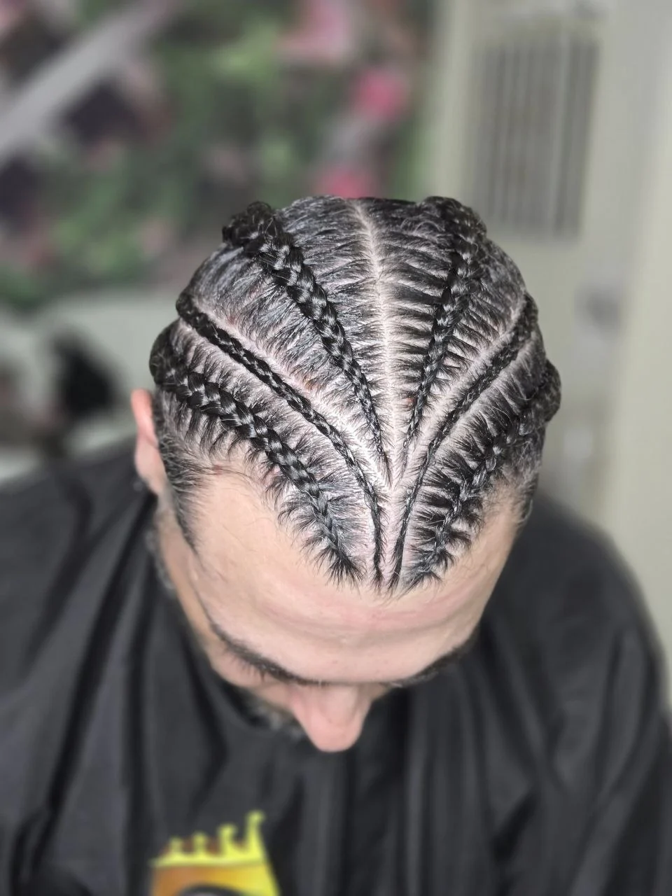 Top-down view of a man's head with sleek cornrow braids arranged in a fanned, star-like pattern radiating from the crown.